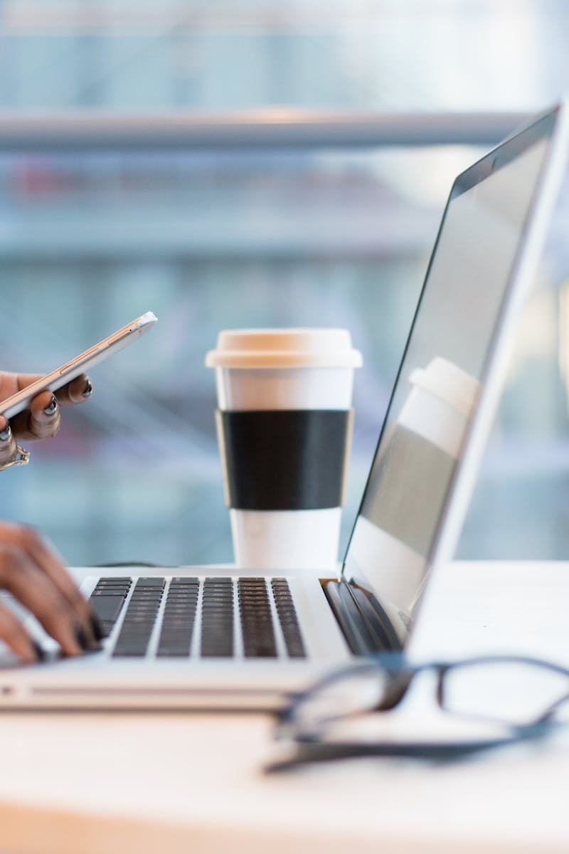 Close-up of hands using a laptop and phone with coffee on a modern office desk.